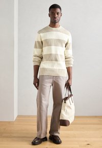 Helle beige, gerippte Sweatshirt mit horizontalen Streifen, kombiniert mit hellkhakifarbenen Hosen und einer Stofftragetasche mit braunen Akzenten. Braune Schuhe.