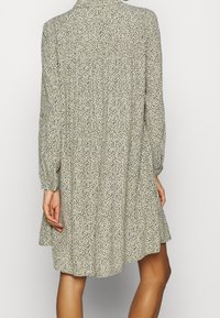 Pitkähihainen mekko vaaleasta beigestä kankaasta, jossa on hajaantunut musta ja tumma vihreä kuvio, pleatattu selkä ja väljä istuvuus.