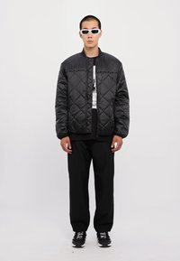 Black quilted puffer jacket with diamond stitching, paired with a graphic black T-shirt and loose black pants. Accessories include white sunglasses.