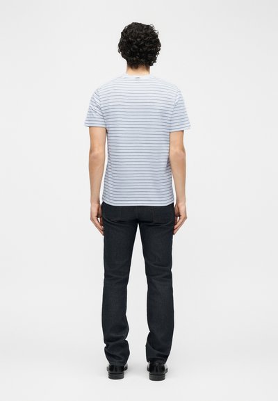 Άνδρας με σγουρά σκούρα μαλλιά, φορώντας ανοιχτό μπλε ριγέ μπλουζάκι, σκούρο τζιν και μαύρα παπούτσια, στέκεται γυρισμένος με την πλάτη σε άσπρο φόντο.