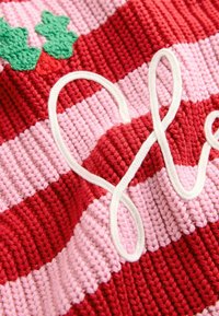 Gebreide stof met gedurfde rode en roze strepen, verfraaid met groene bladeren en een witte, gekrulde decoratieve element.