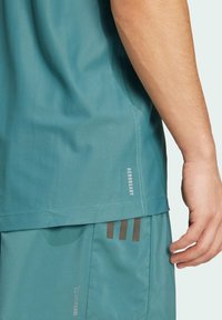 T-shirt de sport bleu-vert avec un tissu texturé, comportant une étiquette imprimée "AEROREADY" sur le côté et un design sans coutures. Shorts coordonnés visibles en bas.