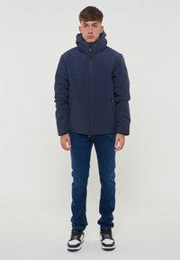 Veste imperméable navy avec un col haut, fermeture éclair sur le devant, deux poches latérales et une coupe ajustée, portée sur un jean bleu et des baskets noires.