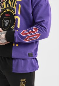 Felpa viola con dettagli neri, caratterizzata da grafiche audaci gialle e rosse, un grande '22' sulla manica e un'etichetta del logo del brand dettagliata.