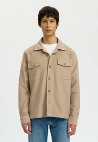 Jonge man in een beige knopenjasje met twee borstzakken over een wit shirt en blauwe spijkerbroek, staand tegen een plain witte achtergrond.