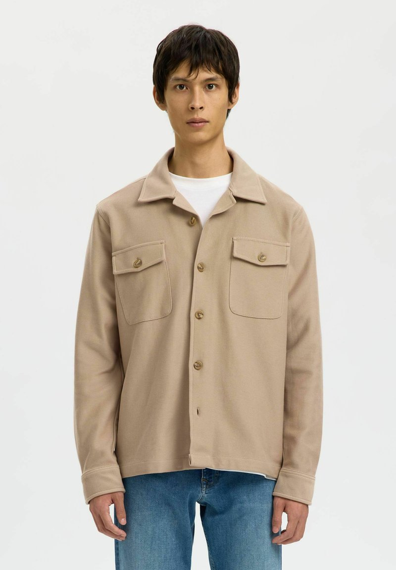 Jonge man in een beige knopenjasje met twee borstzakken over een wit shirt en blauwe spijkerbroek, staand tegen een plain witte achtergrond.