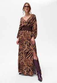 Długa brązowa sukienka w zebrę, z dekoltem w kształcie litery V, długimi rękawami i paskiem w talii. Posiada boczne rozcięcie i jest zestawiona z burgundowymi bootami.