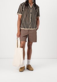 Korte mouwen overhemd met zwart en beige strepen, gecombineerd met een bruine short. Een crèmekleurige handtas en beige sneakers maken de outfit compleet.