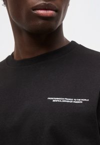 Κοντινό πλάνο ενός ατόμου που φοράει μαύρο πουκάμισο με λευκό κείμενο στο στήθος που γράφει "Από το Farmácia Franco στον κόσμο Benfica, εμπνευσμένο από πάθος."