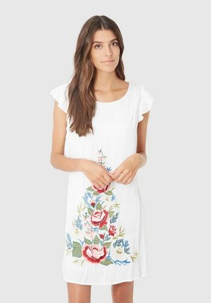 Joven mujer con cabello largo y castaño, vistiendo un vestido blanco con bordados florales en rojo, rosa y azul, de pie frente a un fondo liso.