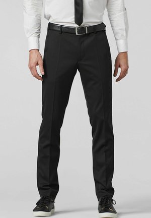 Man draagt zwarte nette broek, zwarte riem, zwarte das, wit overhemd en zwarte sneakers, staand tegen een effen achtergrond.