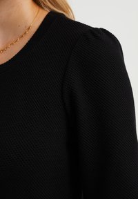 Svart strukturerad stickad tröja med rund halsringning och puffärm. Inkluderar ett halsband i guldton.