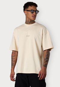 Beige Baumwoll-T-Shirt mit Rundhalsausschnitt und kurzen Ärmeln. Verfügt über ein kleines gesticktes Logo auf der Brust. Getragen mit dunklen Cargo-Hosen.