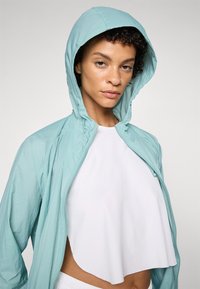 Veste zippée bleu clair avec capuche, fabriquée à partir d'un matériau léger. Présente un petit logo sur la manche droite et un top blanc court en dessous.