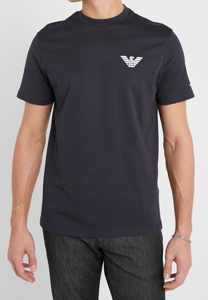 T-shirt en coton bleu marine avec col rond, manches courtes et un logo blanc sur la poitrine gauche. Texture lisse et coupe ajustée.