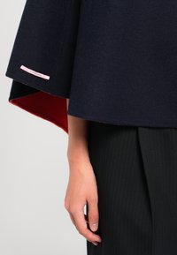 Poncho en laine bleu marine avec doublure rouge, présentant un ourlet incurvé et une étiquette rose. Main visible tenant le bord, stylisé avec un pantalon à rayures.