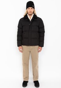 Sort pufferjakke med krave af falsk pels, med knaplukning og to lommer, båret over en lys skjorte og beige bukser. Sort beanie.