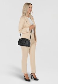 Borsa a mano in pelle nera con hardware dorato, forma ovale, con doppio manico e una lunga tracolla regolabile, esposta accanto a un abito beige.