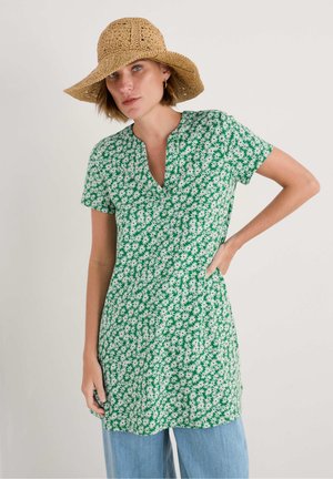Vestido verde de túnica con estampado floral blanco, mangas cortas y escote en V. Combinado con jeans de pierna ancha en azul claro y un sombrero de paja.