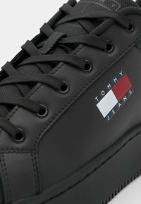 Zapatilla negra con parte superior de cuero texturizado, puntera redonda y cordones negros. Presenta un pequeño logotipo "TOMMY JEANS" en azul marino, rojo y blanco en el costado.
