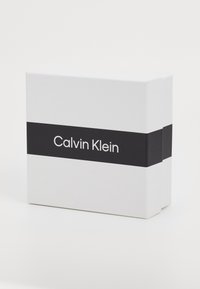 Valge ristkülikukujuline kast musta ribaga, millel on valge tekst "Calvin Klein." Sile pind ja minimaalne disain.