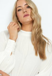 Hvid tekstureret bluse med høj hals, plisseret design og lange puffærmer, som har knapdetaljer ved manchetterne.