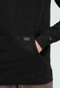 Sudadera negra con un bolsillo frontal, puños y dobladillo acanalados. Presenta una pequeña etiqueta en el lado inferior con "SOLID" y "Desde 1989".