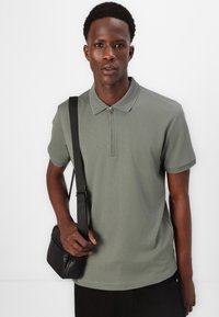 Kortermet polo skjorte i olivengrønn, med glidelås hals og strukturert krage. Brukt med en svart crossover-veske.