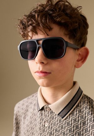 Boy with curly hair wearing large black sunglasses and a beige patterned collared shirt, facing forward with neutral expression.