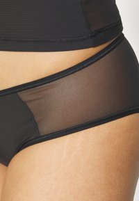 Black bikini bottom featuring a sheer panel on the side, with smooth, stretchy fabric and a seamless finish.