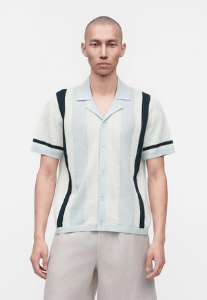 Homme au crâne rasé portant une chemise en maille à manches courtes avec des rayures verticales bleu clair, blanc et bleu marine, et un pantalon clair.