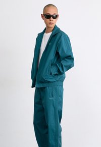 Ensemble de survêtement en teal fabriqué dans un tissu léger, comprenant une veste zippée et des poignets élastiques, associé à un pantalon assorti et des accents de logo subtils.