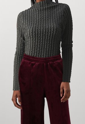 Personne portant un haut à manches longues gris texturé et un pantalon en velours bordeaux taille haute, debout devant un fond clair uni.