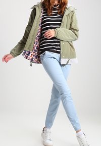 Ljust grön huva jacka med ett färgglatt mönstrat foder, randig svart och vit skjorta, ljusblå byxor och vita sneakers.