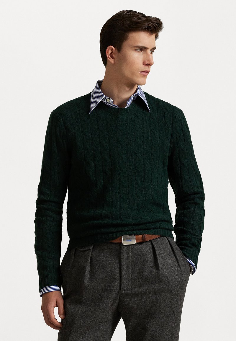 Un pull en tricot torsadé vert foncé avec un col rond, superposé à une chemise à col rayée bleu clair et blanche, accompagné de pantalons gris.