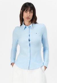 Camicia a maniche lunghe a strisce blu chiaro con colletto a bottoni, patch logo discreto e design aderente. Il tessuto ha una consistenza liscia.
