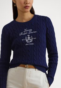 Temno modra pulover z vizo v obliki mrežastih pletenin, z okroglim izrezom in vezenim logotipom. Kombiniran s svetlejšimi hlačami in rjavim pasom z zlato zaponko.