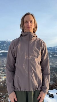 Άνδρας με μαλλιά μέχρι τους ώμους, ντυμένος με γκρι μπουφάν Salomon, στέκεται έξω με χιονισμένα βουνά και μια πόλη στο φόντο.