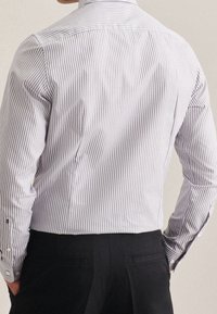 Uomo che indossa una camicia bianca a maniche lunghe aderente con sottili righe grigie e pantaloni neri, in piedi con le mani in tasca, di spalle rispetto all'osservatore.