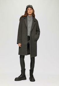 Grijze wolcoat met een notch-kraag, gecombineerd met een lichtgrijze coltrui en zwarte leren legging-achtige broeken. Zwarte veterschoenen voor in de strijd maken de look compleet.
