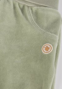 Green corduroy pants with a soft texture. Features a round, tan logo patch on the left pocket. Elastic waistband at the top.