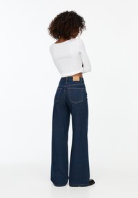 Mujer con cabello rizado que lleva una camiseta de manga larga blanca y pantalones de mezclilla anchos de color azul oscuro, de pie con los brazos cruzados, mirando hacia el lado opuesto.