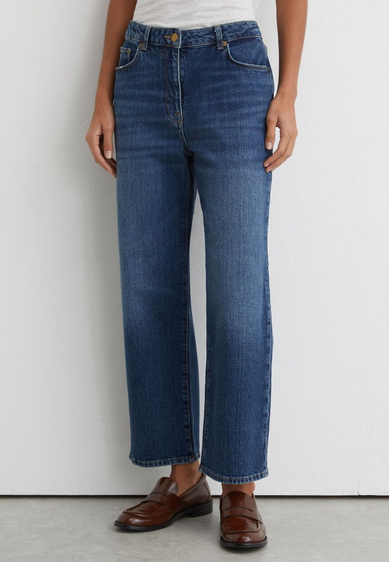 Person iført højtaljede blå jeans med lige ben og brune læder loafers, stående mod en ensfarvet hvid væg.