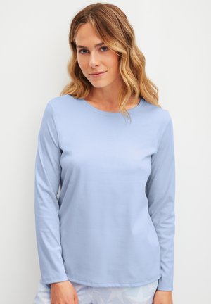 Femme aux cheveux blonds ondulés portant une chemise à manches longues bleu clair, debout devant un fond blanc uni, faisant face à l'avant.