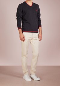 Dunkelgrauer V-Ausschnitt-Pullover mit roten Akzenten, cremefarbene Hose und weißeSneaker. Der Pullover hat lange Ärmel und eine figurbetonte Passform.
