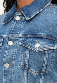 Denim jakna u svijetloplavoj boji s klasičnom ovratnikom, srebrnim dugmadi, dva prednja džepa i strazom s rhinestone detaljima na ramenima.