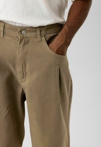 Kaki katoenen broek met een relaxte pasvorm, voorzien van zijzakken en een voorplooi. De tailleband heeft een knoopsluiting en riemlussen.