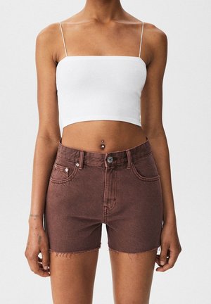 Kvinde iført en hvid kort spaghetti-strop top og højtaljede brune denimshorts med rå kanter, stående mod en ensfarvet baggrund.