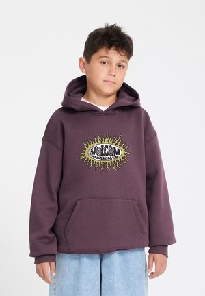 Garçon aux cheveux courts et foncés portant un sweat à capuche violet surdimensionné avec un graphique de soleil jaune et un jean bleu clair, debout devant un fond blanc.