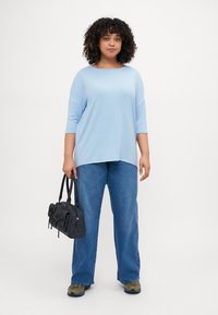 Light blue, loose-fit top with three-quarter sleeves paired with blue jeans. The model holds a black handbag with bow accents.